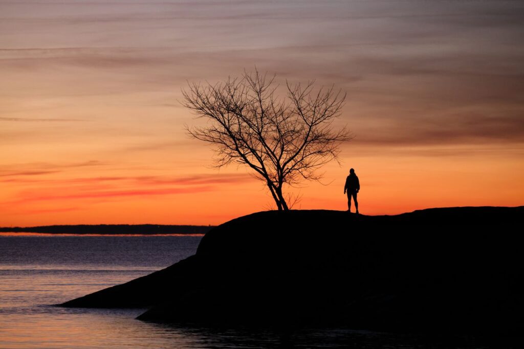A lone figure stands on a rocky shore, silhouetted against a vibrant sunset over the sea.
