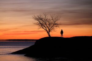 A lone figure stands on a rocky shore, silhouetted against a vibrant sunset over the sea.